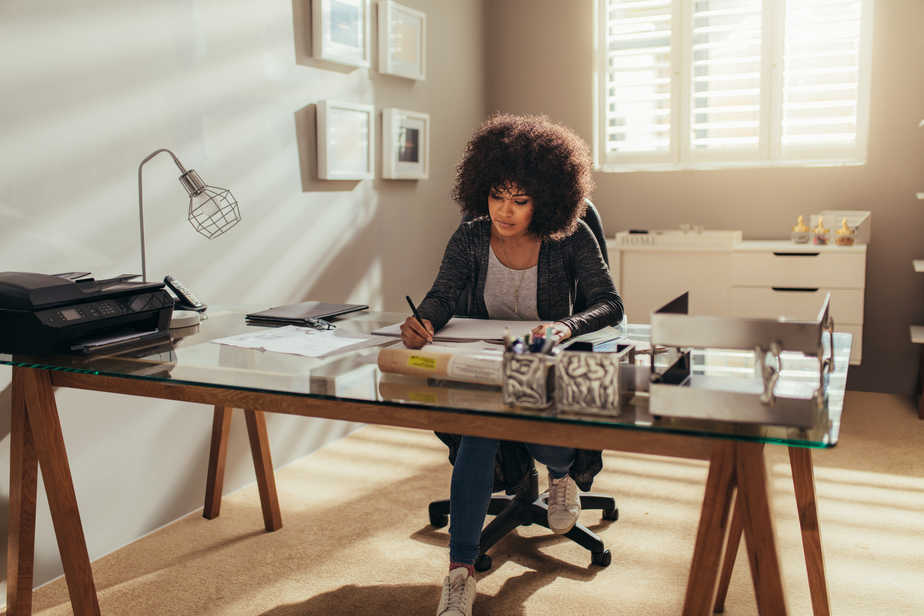 A woman working in a home office at a glass top desk. She's writing with a pen and has a printer and lamp on the desk.