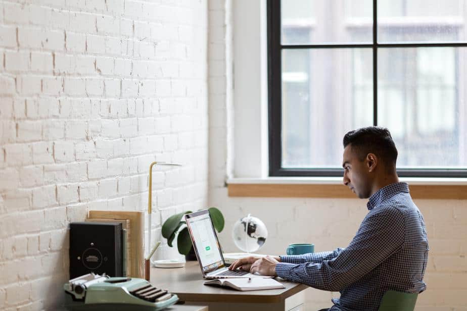 A man in a blue checkered shirt sits at a desk working from home on his laptop, and a typewriter to his left.