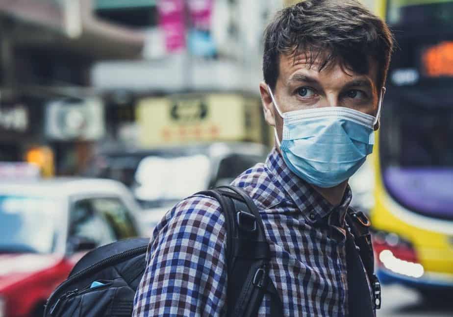 A man wearing a surgical mask and a plaid shirt, in front of city traffic.