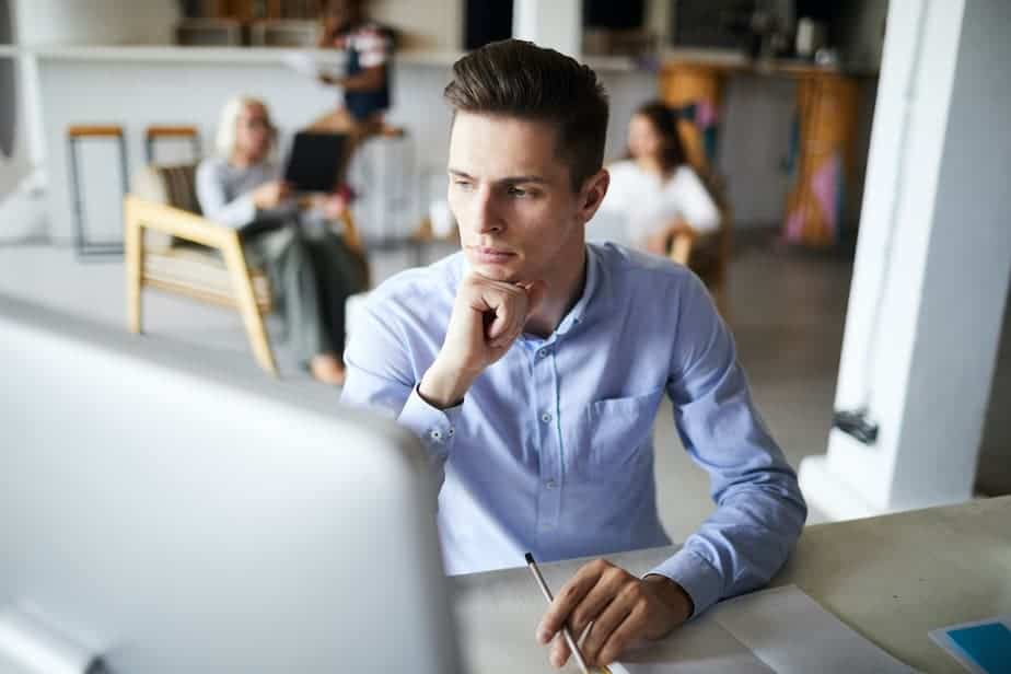 A man sits at a desk, holding a pencil and looking at a monitor. Behind him are 2 women sitting in chairs working on laptops.