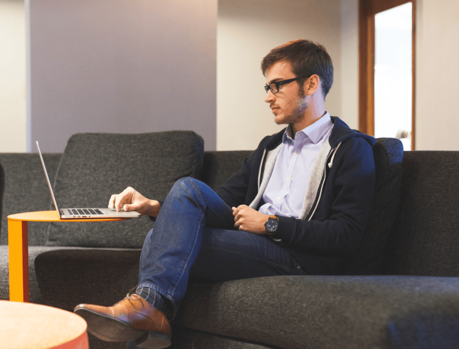 A man wearing a jacket, jeans and brown shoes, sitting on a couch in a waiting room while working on his laptop.