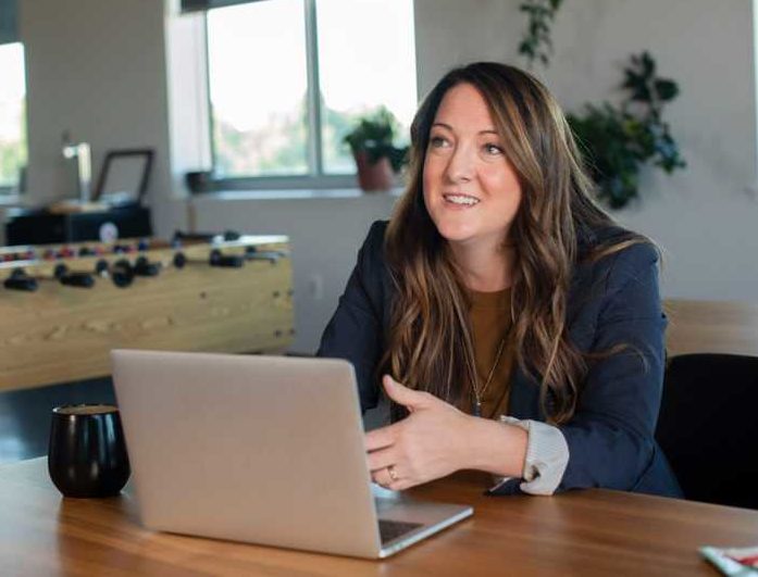 A woman sitting at a table with a laptop and coffee mug in front of her. She is looking up as if listening to someone.