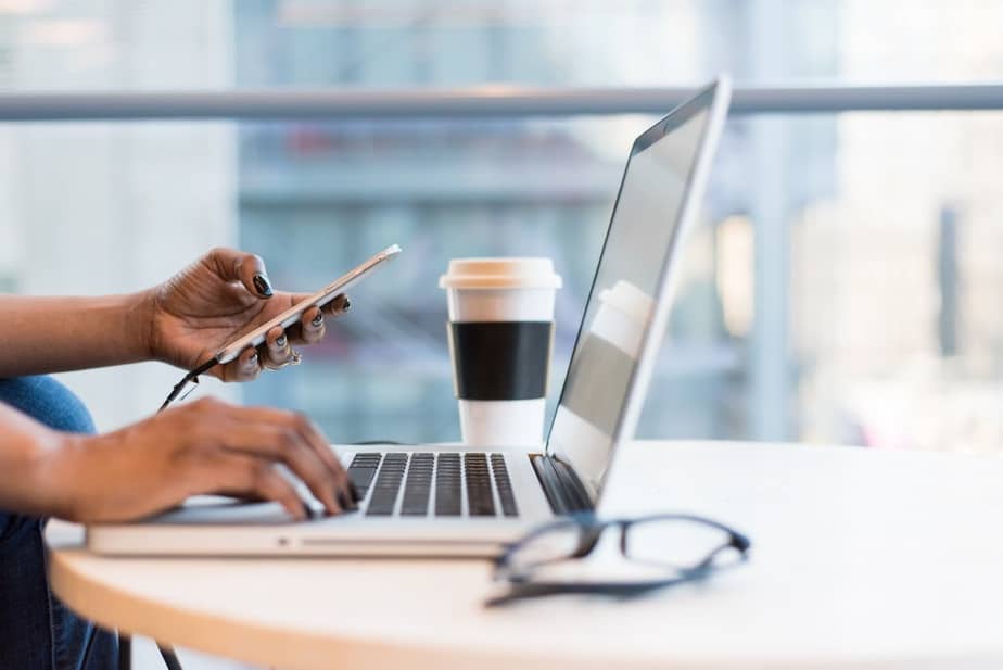 A woman holds a phone in her left hand and types on a laptop with the other. There's a coffee cup and pair of glasses.