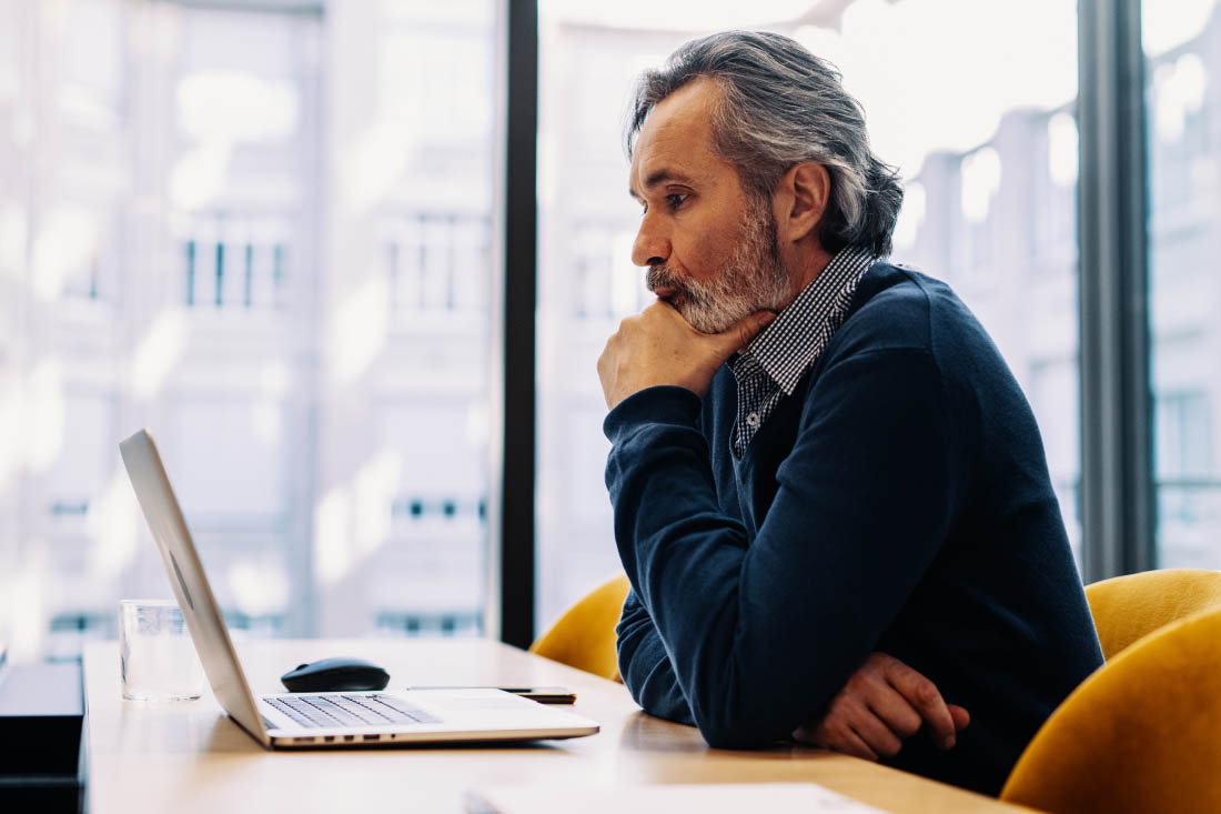 Middle-aged man working remotely on a laptop in a bright office space, reflecting deeply.